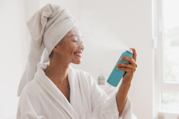 Jovem vestindo roupão e toalha de cabeça usando spray de oxigênio de atualização facial após o tratamento em um salão de spa de beleza.