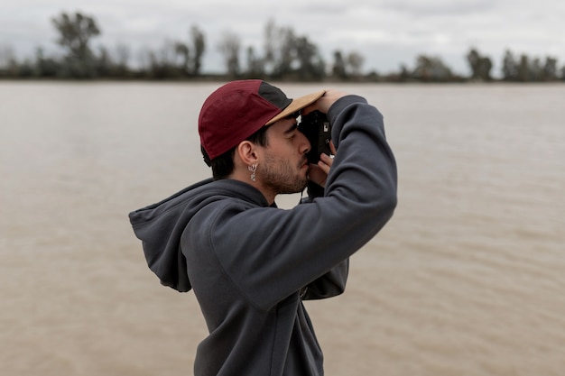 Jovem vestindo um suéter cinza e boné vermelho tirando fotos em um píer próximo a um rio