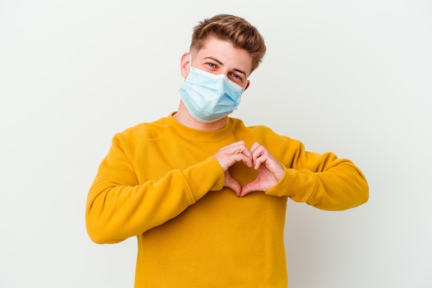 Jovem vestindo uma máscara para coronavírus isolada na parede branca, sorrindo e mostrando um formato de coração com as mãos