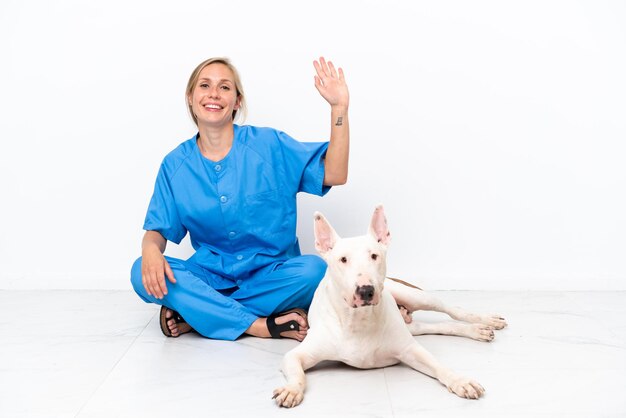 Jovem veterinária inglesa sentada no chão com um cachorro saudando com a mão com uma expressão feliz