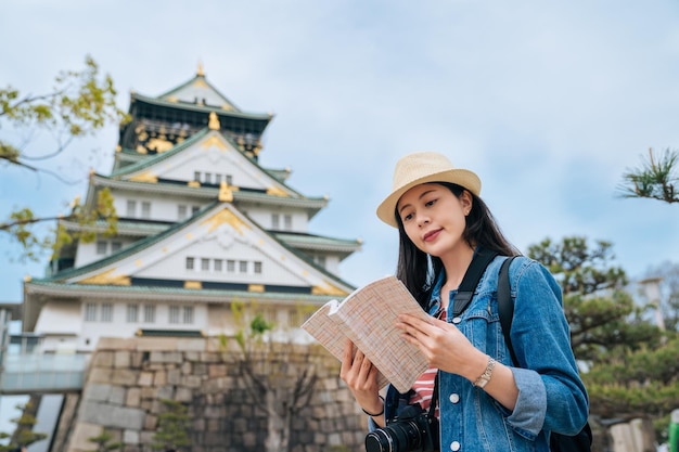 jovem viajante asiático segurando o guia visitando o castelo histórico de osaka com céu azul claro ao fundo. guia de leitura turística feminina e encontrar o preço da entrada. conceito de auto viagem jp.