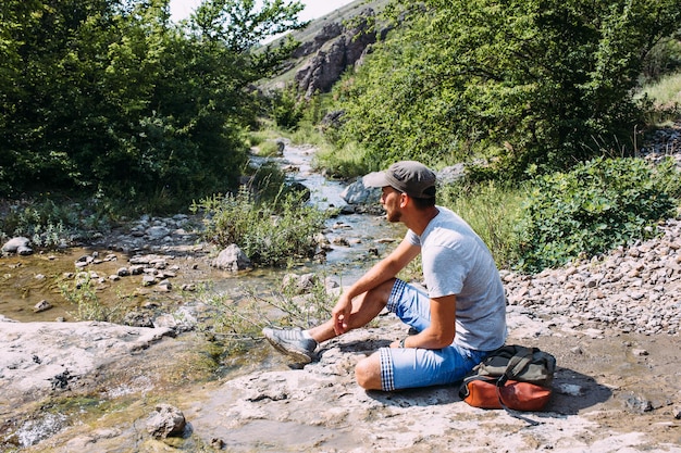 Jovem viajante caminhando sozinho à beira de um rio de montanha com paisagem rochosa de verão