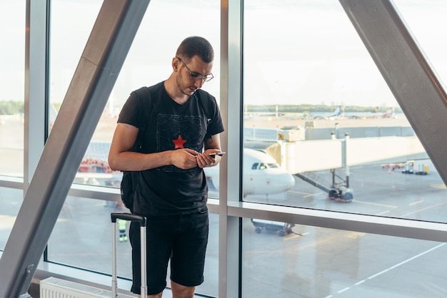 Jovem viajante casual masculino no aeroporto segurando telefone inteligente perto de janelas de portão em aviões na pista