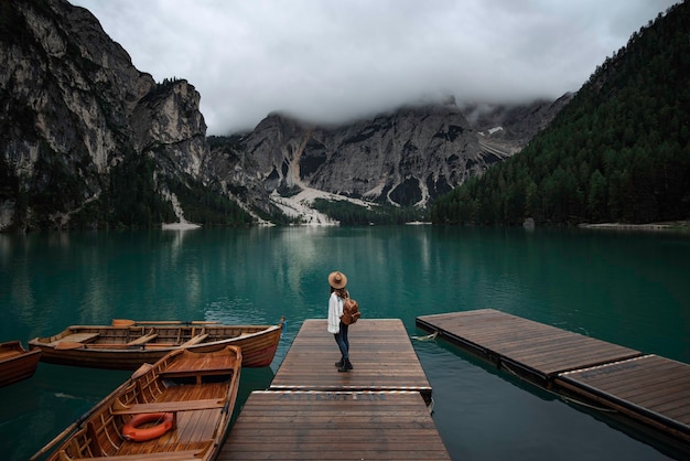 Jovem viajante com chapéu e mochila vintage no cais do lago di Braies, cercada pelas montanhas dos Alpes italianos