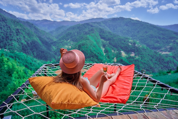 Jovem viajante com chapéu para relaxar com uma taça de vinho e deitada em uma rede durante o momento de vida feliz e livre, contra o fundo de grandes montanhas verdes