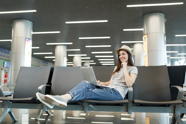 Jovem viajante encantadora mulher de chapéu trabalhando em um laptop enquanto espera no saguão do aeroporto internacional