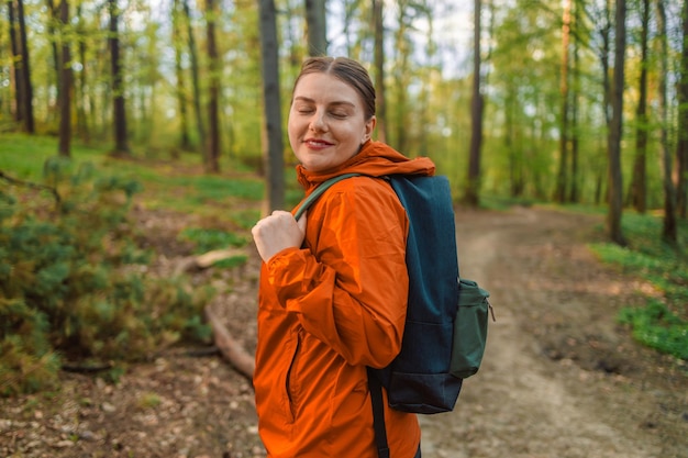 Jovem viajante feliz em jaqueta laranja com mochila na floresta de primavera curtindo caminhar respirando