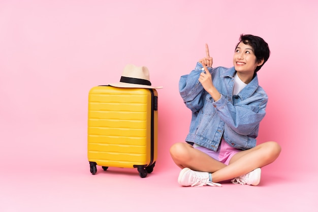 Jovem viajante mulher vietnamita com mala sentada no chão parede rosa apontando com o dedo indicador uma ótima idéia