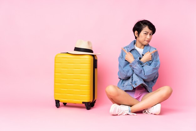 Jovem viajante mulher vietnamita com mala sentado no chão parede rosa apontando para os lados com dúvidas