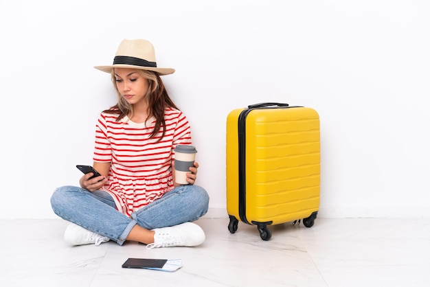Jovem viajante sentada no chão, isolada no fundo branco, segurando um café para levar e um celular