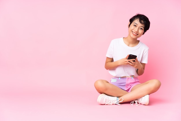 Jovem vietnamita com cabelo curto, sentada no chão sobre parede rosa isolada, enviando uma mensagem com o celular