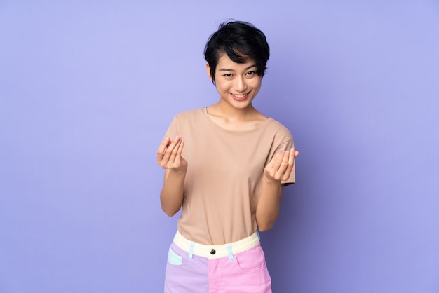 Jovem vietnamita com cabelo curto sobre parede roxa isolada, fazendo gesto de dinheiro