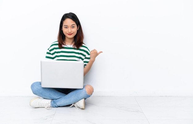 Jovem vietnamita com um laptop sentada no chão, isolada na parede branca, apontando para o lado para apresentar um produto