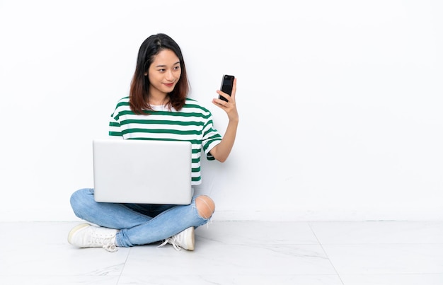 Jovem vietnamita com um laptop sentada no chão, isolada na parede branca fazendo uma selfie