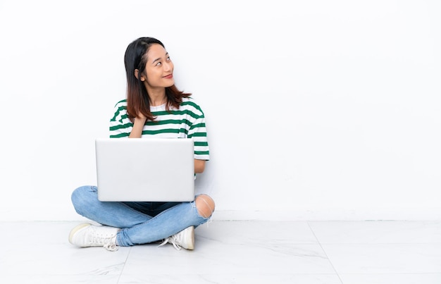 Jovem vietnamita com um laptop sentada no chão, isolada na parede branca, pensando em uma ideia