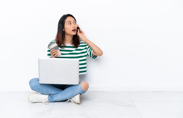 Jovem vietnamita com um laptop sentada no chão, isolada na parede branca, segurando um café para levar e um celular