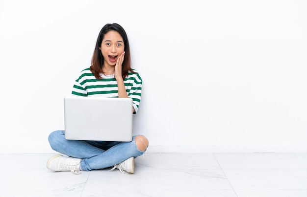 Jovem vietnamita com um laptop sentado no chão isolado na parede branca com surpresa e expressão facial chocada