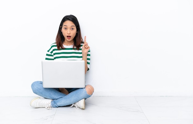 Jovem vietnamita com um laptop sentado no chão isolado na parede branca pensando em uma ideia apontando o dedo para cima