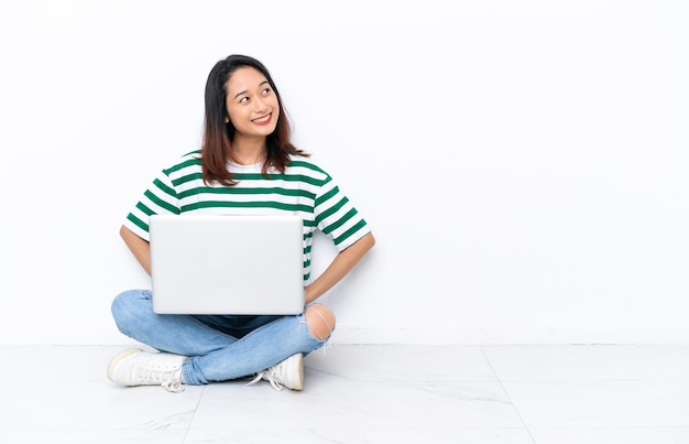 Jovem vietnamita com um laptop sentado no chão isolado na parede branca posando com os braços no quadril e sorrindo