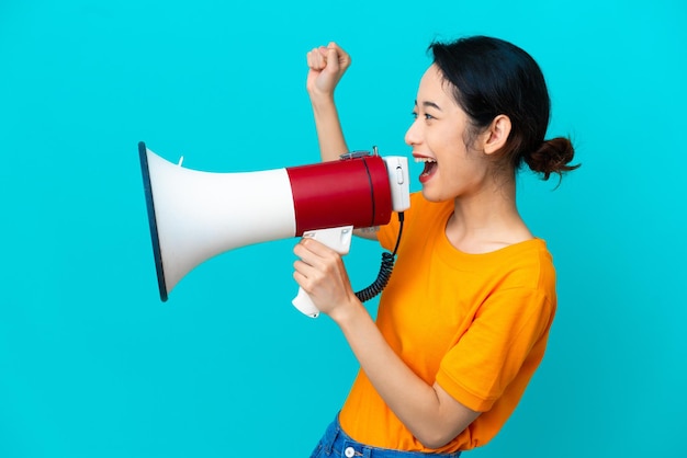 Jovem vietnamita isolada em fundo azul gritando através de um megafone para anunciar algo em posição lateral