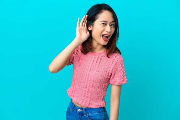 Jovem vietnamita isolada em fundo azul ouvindo algo colocando a mão na orelha