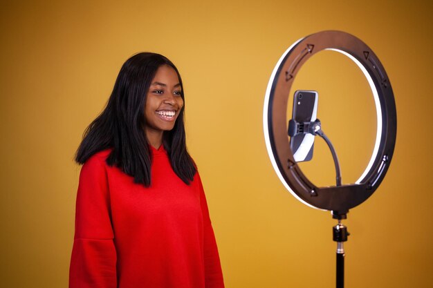 Jovem vlogger afro-americana sorridente em um capuz vermelho com uma lâmpada de anel iluminada e telefone