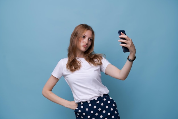 Jovem vlogger com cara de surpresa tira foto de si mesma no telefone enquanto transmite em um fundo azul em uma camiseta branca Menina bonita caucasiana Selfie concept story and blog