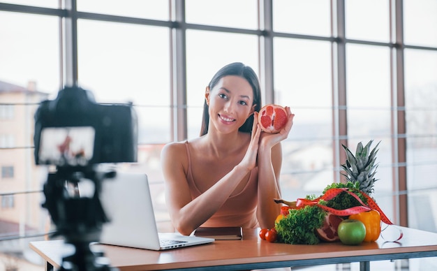 Jovem vlogger de fitness fazendo vídeo dentro de casa sentado perto da mesa com comida saudável.