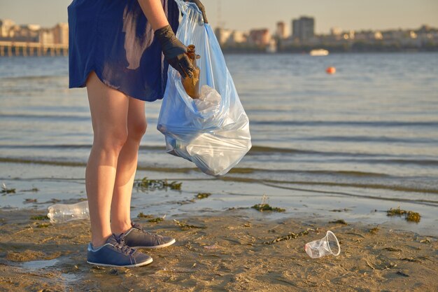 Foto jovem voluntária de luvas pretas caminha com saco de lixo em uma praia suja do rio