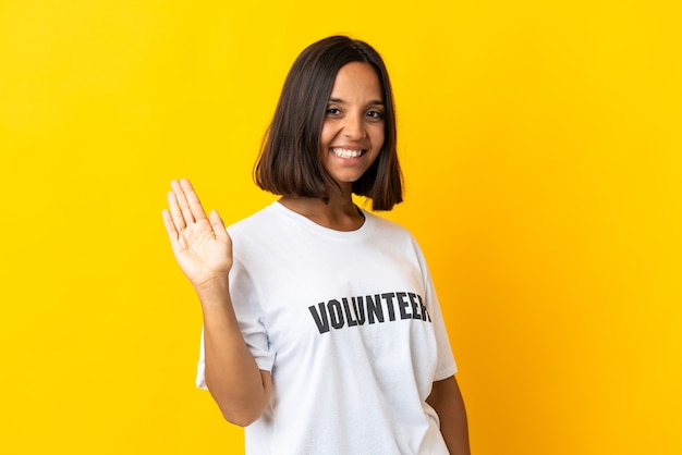 Jovem voluntária isolada em fundo amarelo saudando com a mão com expressão feliz