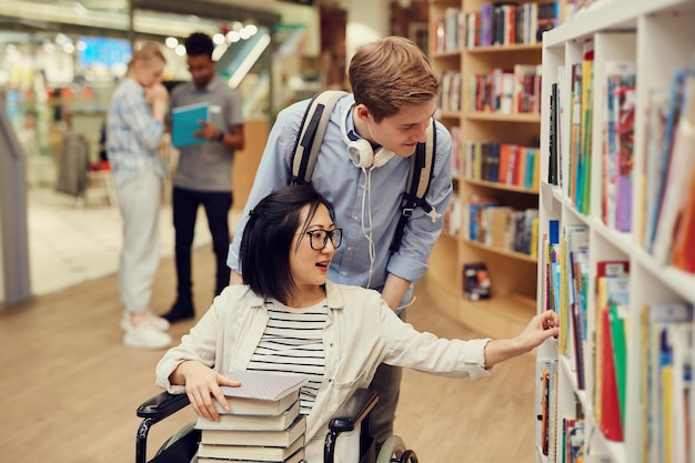 Jovem voluntário cuidadoso empurrando cadeira de rodas com uma estudante asiática enquanto a ajudava na biblioteca
