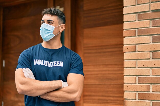 Jovem voluntário hispânico com os braços cruzados usando máscara médica na cidade.