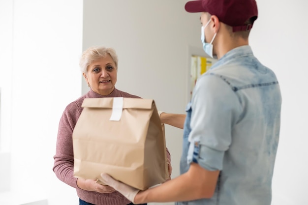 Jovem voluntário masculino com máscara dá caixas a uma mulher idosa com comida perto de sua casa. Filho homem ajuda uma mãe idosa solteira. Apoio familiar, carinho. Em quarentena, isolado. Coronavírus (COVID-19. Doação