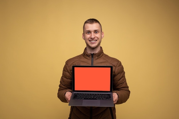 Joven de 20 años con chaqueta marrón, con fondo amarillo, usa trabajo portátil, usa trabajo portátil con espacio de trabajo de pantalla en blanco