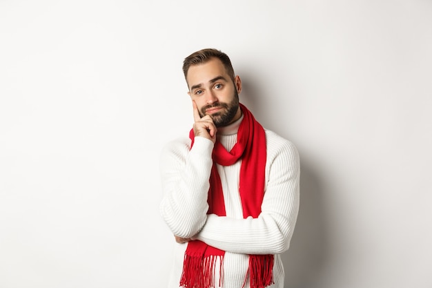 Joven aburrido mirando indiferente y cansado a la cámara, escuchando a alguien, de pie en suéter y bufanda de invierno, fondo blanco.