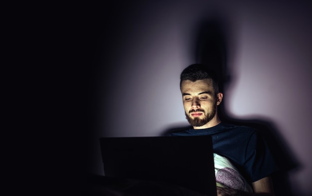 Joven acostado en la cama y mirar la pantalla del portátil. Tranquilo chico concentrado trabajando horas extras jugando o entreteniéndose. Sala de noche.