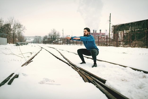 Joven activo agachado y haciendo ejercicios en el lugar público entre el viejo ferrocarril durante el entrenamiento de invierno afuera. Copie el espacio.