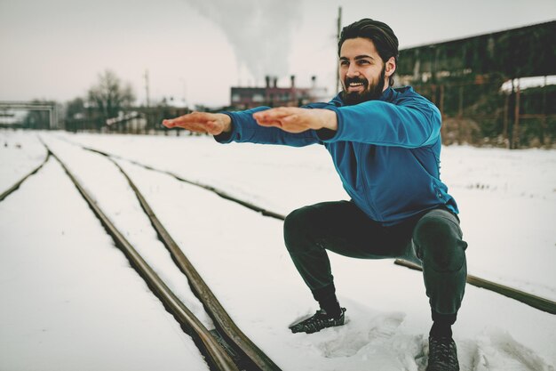 Joven activo agachado y haciendo ejercicios en el lugar público entre el viejo ferrocarril durante el entrenamiento de invierno afuera. Copie el espacio.