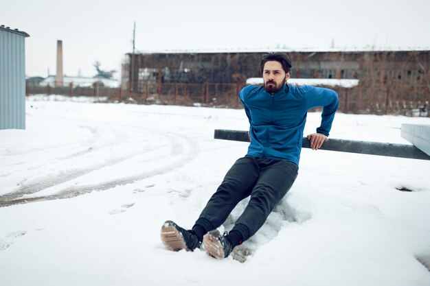 Joven activo estirándose y haciendo ejercicios en el lugar público entre el antiguo ferrocarril durante el entrenamiento de invierno afuera. Copie el espacio.