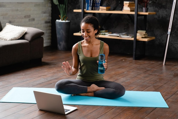 joven adolescente africana mujer con ropa de fitness teniendo videoconferencia en línea en portátil después de entrenar entrenamiento fitness en casa en estera Blogging concepto de vlogging