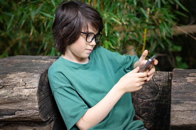 Joven adolescente con anteojos sentado en un banco ecológico en el parque escribiendo un mensaje en su teléfono inteligente usando el teléfono al aire libre en el concepto de redes sociales de verano