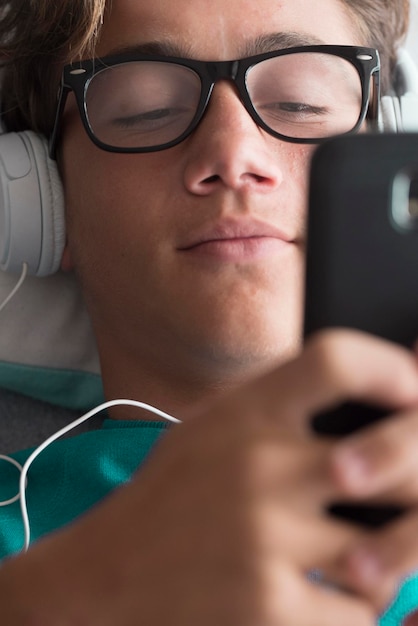 Joven adolescente caucásico guapo usar dispositivo de teléfono celular y escuchar música con auriculares acostarse en casa descargar canciones actividad y concepto de vida conectada moderna