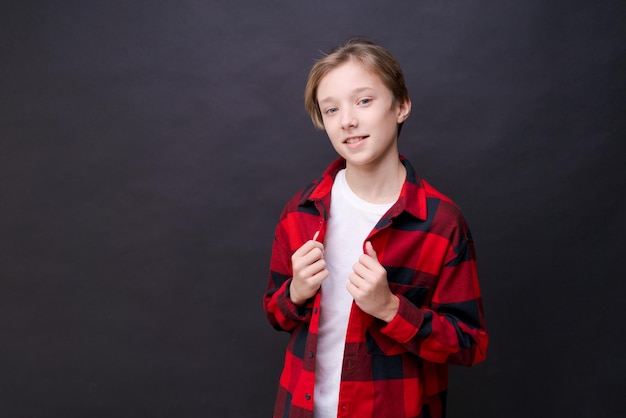 Un joven adolescente caucásico sonriente con dientes divertidos con una simple camiseta casual y una camisa en una jaula roja mirando a la cámara, aislado en un retrato de estudio contra un fondo negro de pared. Concepto de estilo de vida de emociones