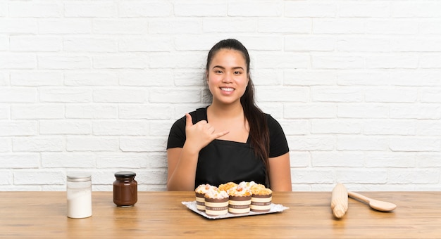 Joven adolescente chica asiática con un montón de pastel de muffin haciendo gesto de teléfono