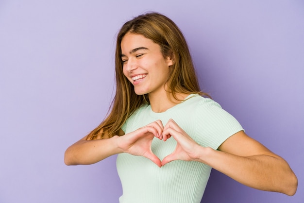 Joven adolescente flaca caucásica sobre fondo púrpura sonriendo y mostrando una forma de corazón con las manos.