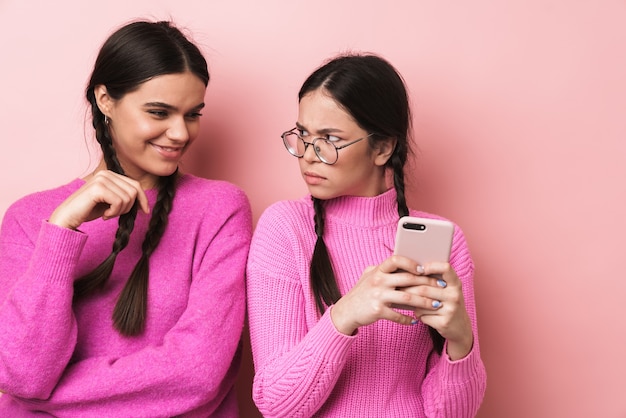 Joven adolescente de pie junto a una amiga feliz y mirando su teléfono celular aislado sobre pared rosa