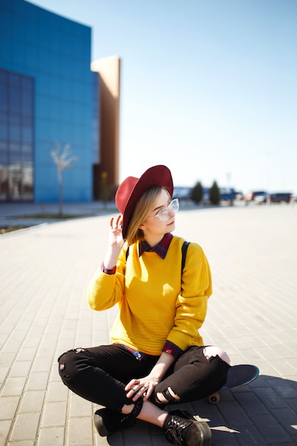 Joven adolescente sentado en una patineta y mensajes de texto en un teléfono móvil. Chica hipster en un suéter amarillo