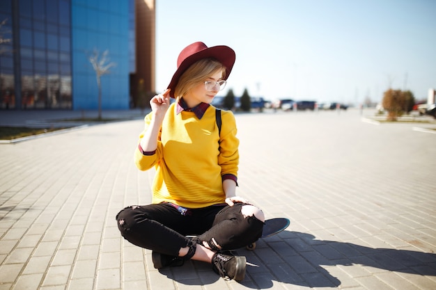 Joven adolescente sentado en una patineta y mensajes de texto en un teléfono móvil. Chica hipster en un suéter amarillo