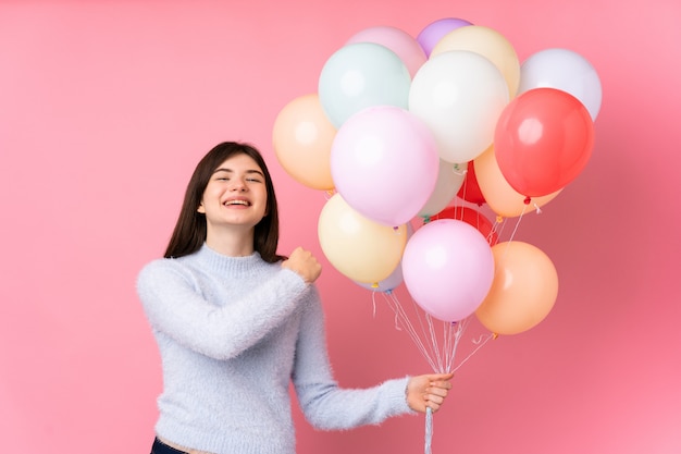 Joven adolescente sosteniendo muchos globos sobre pared rosa celebrando una victoria