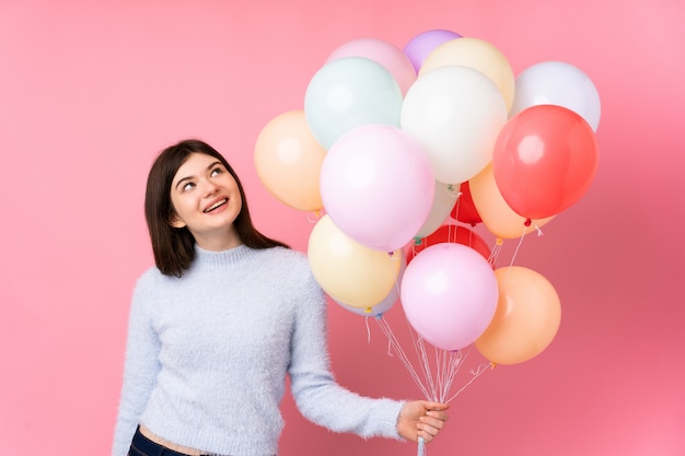 Joven adolescente sosteniendo muchos globos sobre pared rosa riendo y mirando hacia arriba